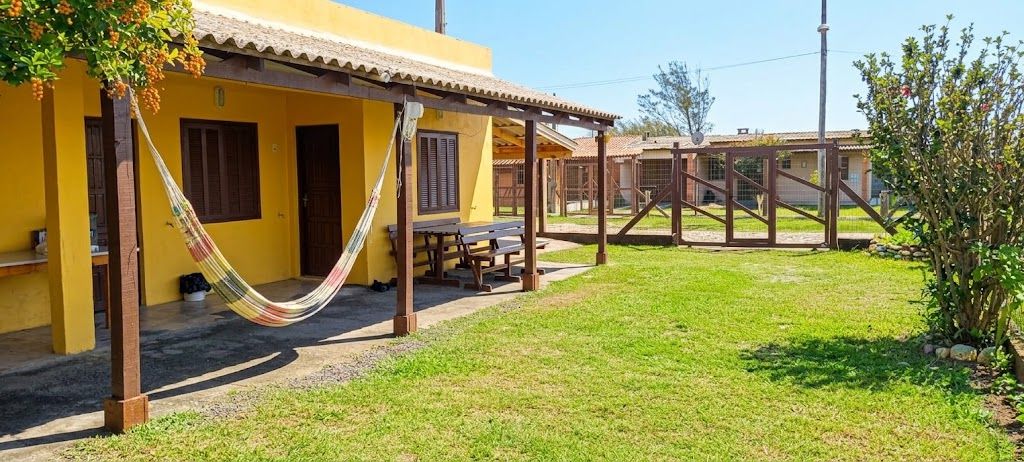 Fachada amarela da Pousada Recanto da Família em Balneário Pinhal, com rede na varanda, mesa de madeira e gramado em dia de céu azul