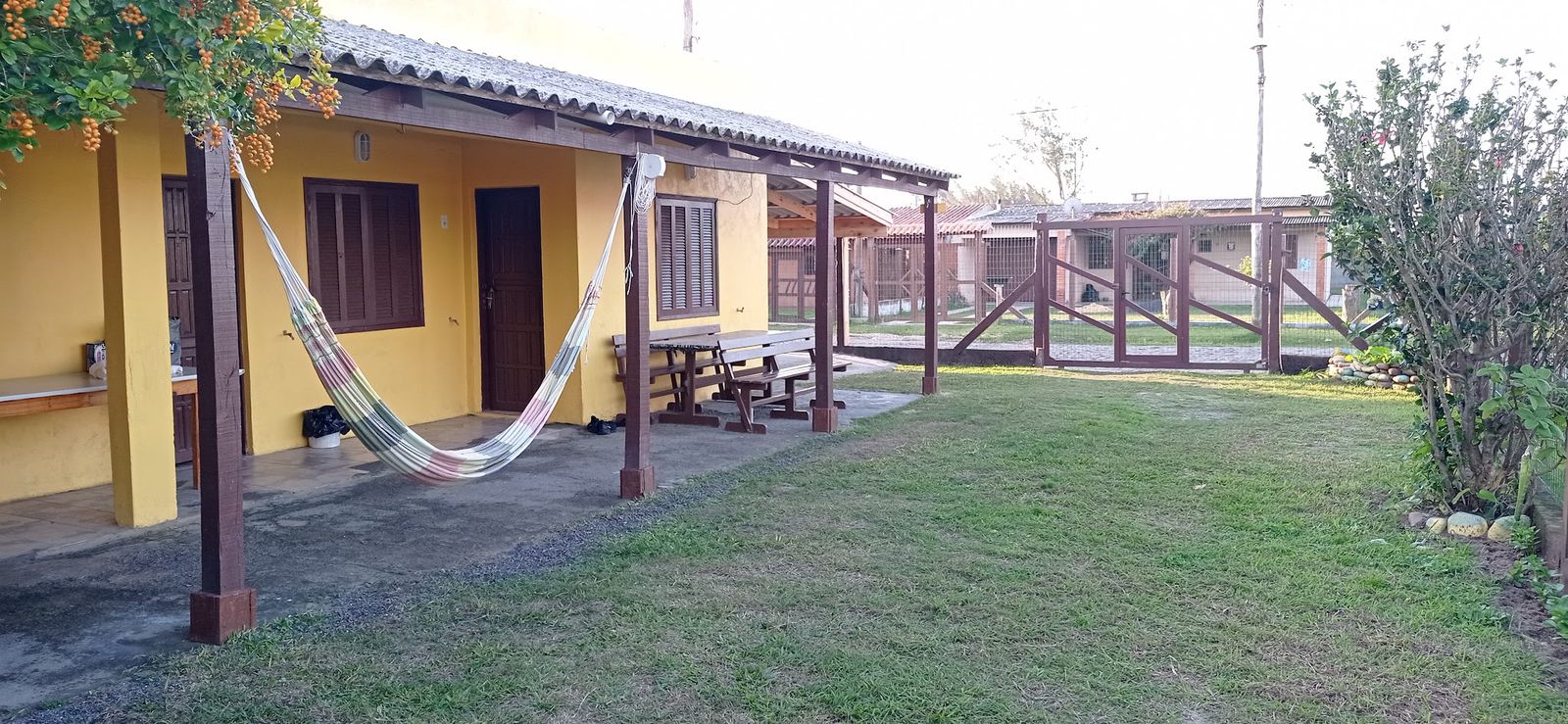Varanda amarela da pousada com rede, mesa e bancos de madeira e gramado, em Balneário Pinhal